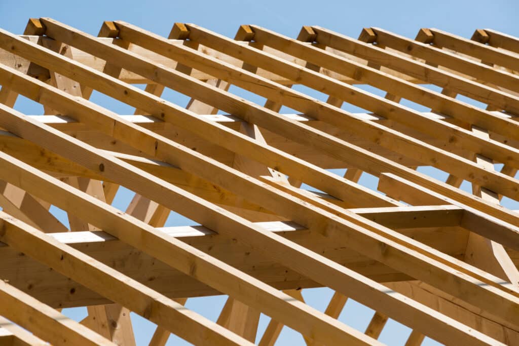 Charpente de toit en bois clair contre un ciel bleu lumineux, montrant la structure d'une maison en construction avec chevrons.