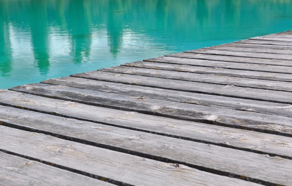 Ponton en bois gris patiné avec des planches marquées, s'avançant sur un lac d'eau turquoise claire et agitée par de légers reflets.